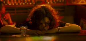 print da cena em que sue lynne, uma mulher branca de cabelos pretos está sentada no bar e a iluminação contém muito vermelho e amarelo